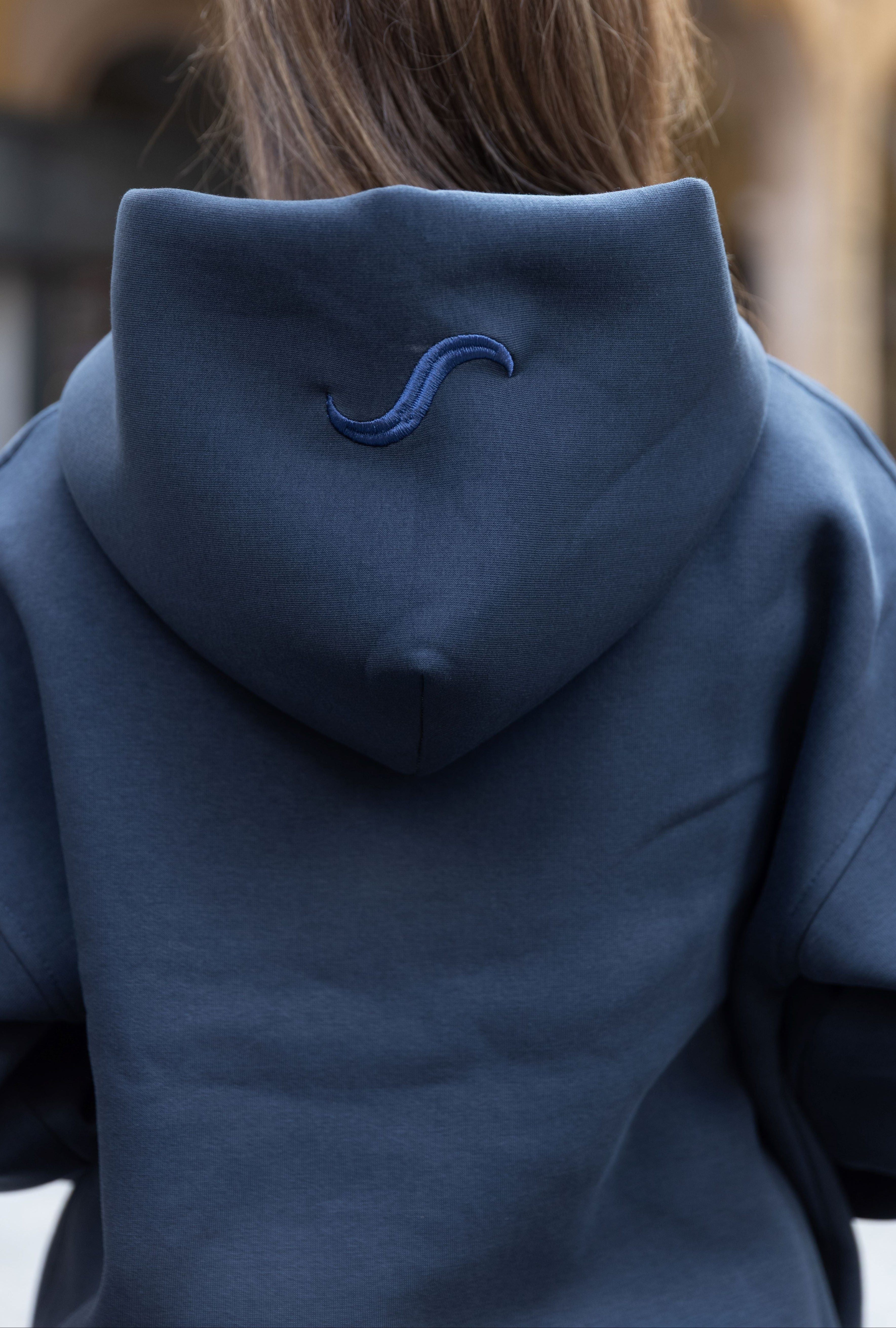Person wearing a dark blue hoodie with a logo on the back, standing outdoors.