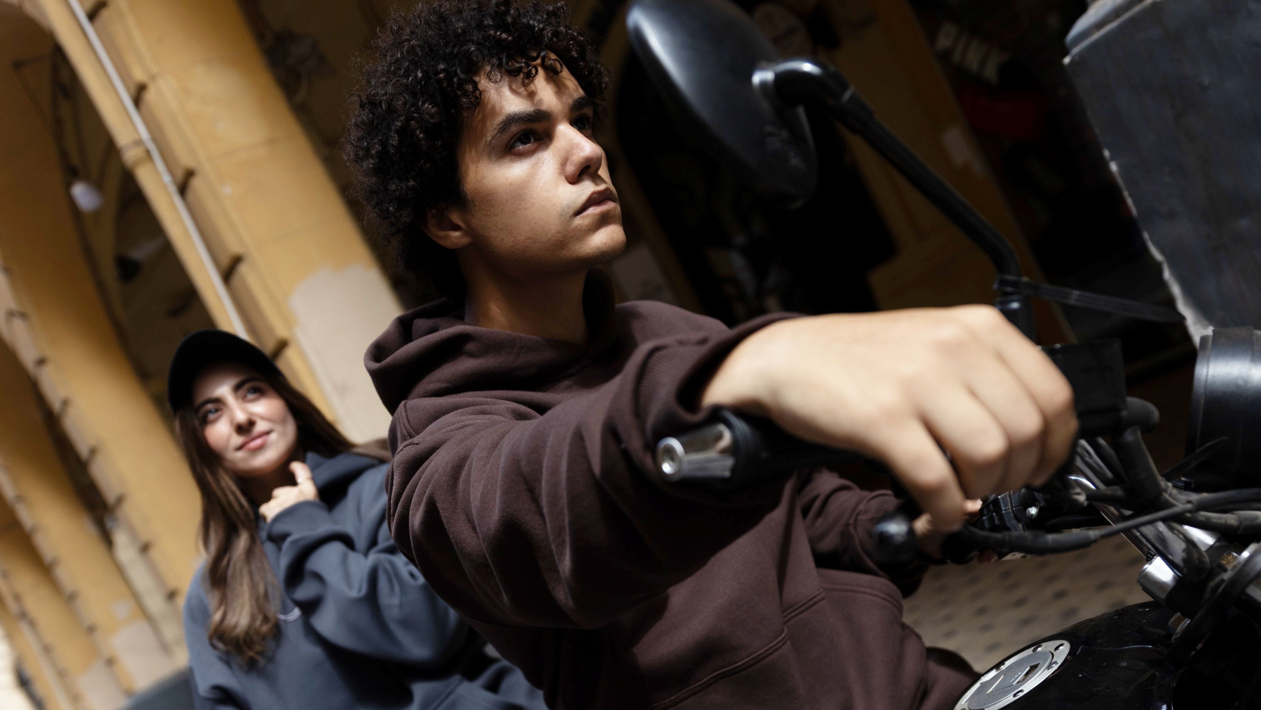 Man sitting on a motorcycle with a woman standing behind him, both wearing dark clothing.